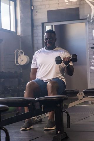 African american man performing seated dumbbell curl on bench in daylight gym interior