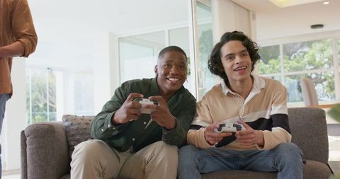 Diverse friends holding controllers playing video games on couch in sunlit living room