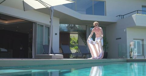 Shirtless man laying out striped towel on luxury pool deck at modern villa under sun