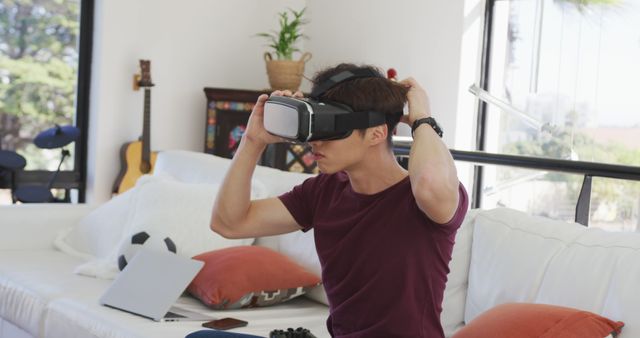 A young man is adjusting his VR headset while sitting on a sofa in a modern and cozy living room. There are pillows, a laptop, and a video game controller nearby, suggesting a relaxed ambiance. Suitable for themes related to virtual reality technology, home entertainment, gaming, and modern lifestyle.