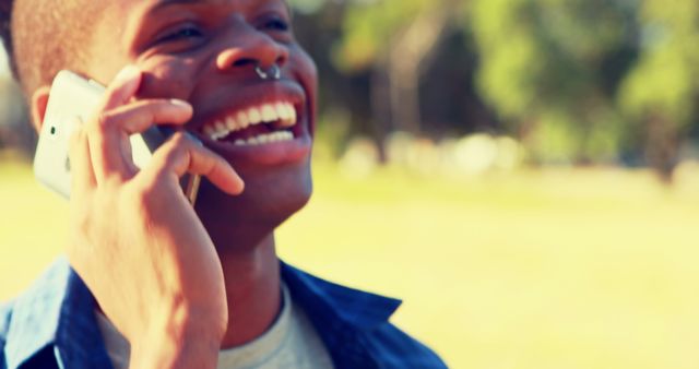 A young man stands outdoors while talking on his smartphone, smiling and looking cheerful. The background is a bright and sunny park, conveying an image of positivity and connectivity. This image is perfect for use in lifestyle blogs, technology advertisements, social media campaigns, articles about communication, or any content showcasing young, happy, and connected individuals enjoying life.