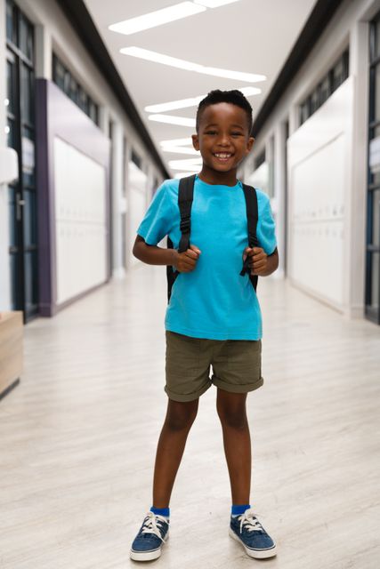 Young African American boy standing in school corridor, smiling and carrying backpack. Ideal for educational materials, back-to-school promotions, and childhood development content.