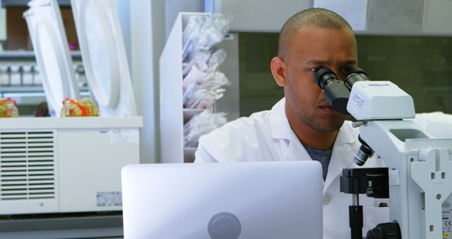 Male scientist in laboratory wearing lab coat, examining specimens through a microscope. Ideal for use in scientific research articles, educational content, medical blogs, or promotional material for pharmaceuticals and biotechnological companies.