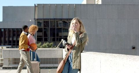 Blonde woman sipping coffee on urban rooftop while diverse friends walk by with basketball