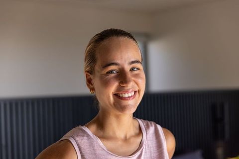 Friendly young woman smiling in cozy bedroom wearing pink sleeveless top and gold earrings