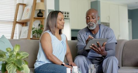 African american nurse reviewing medication with patient on couch using tablet telehealth