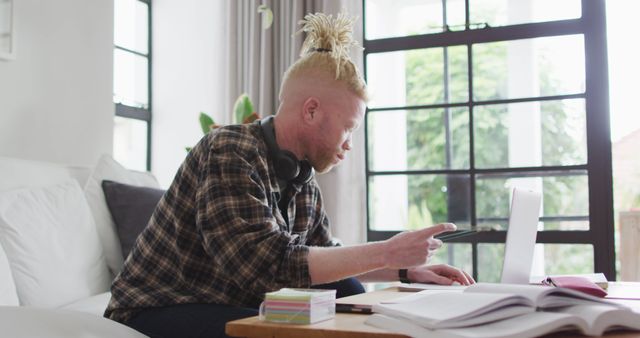 Man working remotely from home while wearing headphones, sitting on couch in modern living room. Ideal for illustrating concepts of work-life balance, remote work, casual work environment, home office setups, and modern living.