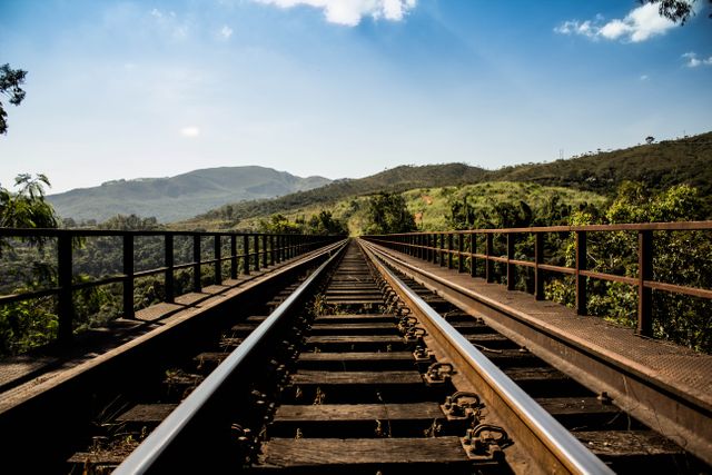 Railroad tracks on a metal bridge stretch across a scenic valley filled with lush greenery and rolling mountains under a bright blue sky. Ideal for themes of travel, adventure, connections, and serenity. Useful for tourism advertising, nature articles, and environmental conservation materials.