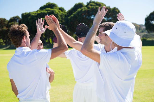 Cricket players in white uniforms are celebrating their victory on a sunny day. They are giving high fives, showing teamwork and joy. This image can be used for sports-related content, team-building promotions, or articles about cricket and outdoor activities.
