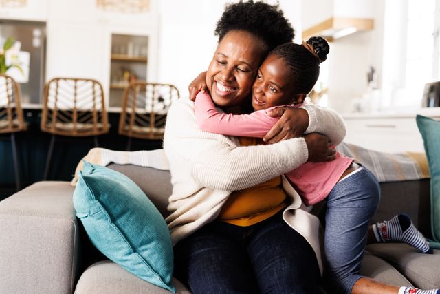 Happy african american granddaughter hugging her smiling grandmother on couch at home. Family, free time and domestic life concept.