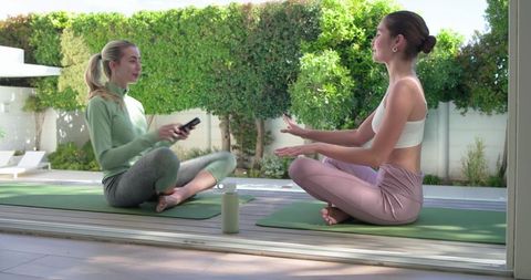 Two women practicing yoga and chatting on sunlit patio deck with water bottle