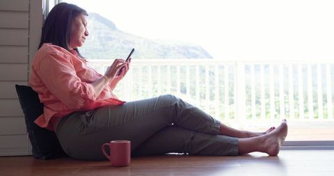 Mature asian woman relaxing on floor by balcony, scrolling smartphone with pink mug