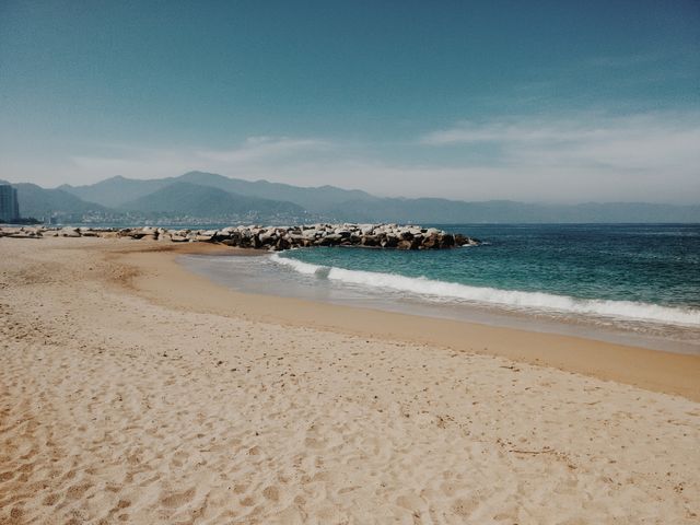 A serene beach with fine golden sand, gentle waves lapping against the shore, and a clear blue sky. The rocky breakwater in the background adds texture to the calm sea, while distant mountains contribute to the picturesque landscape. Ideal for travel blogs, vacation advertisements, nature backgrounds, or web banners promoting seaside destinations and getaways.