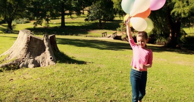 A young girl holds colorful balloons, running through a park with lush green trees and grass on a sunny day. Ideal for content about childhood, outdoor activities, celebrations, and happiness.