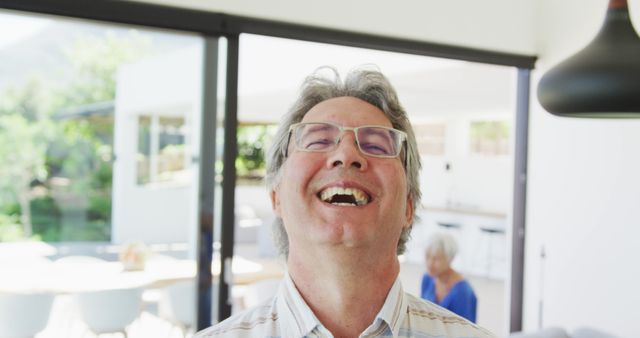 Senior man wearing glasses laughing joyfully, standing indoors in a bright, modern living room. Natural light fills the space, suggesting a warm, inviting atmosphere. Suitable for concepts like happiness in old age, family dynamics, positive emotions, and joyous moments. Great for use in advertisements for senior living, eyewear, lifestyle promotions, and family-oriented content.