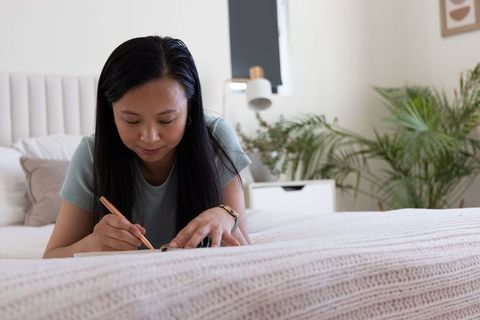 Asian woman lying on bed writing in journal with rose gold pen in minimal bright bedroom