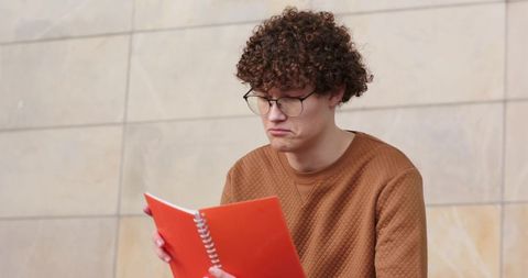 Young man reading orange spiral notebook with thoughtful expression near beige tiled wall