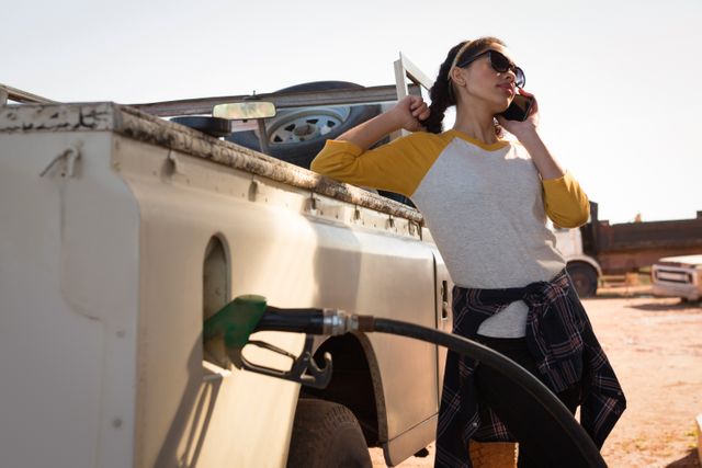 Woman standing next to her car at a gas station, talking on her mobile phone while refueling. She is dressed casually, wearing sunglasses and a plaid shirt tied around her waist. This image can be used for themes related to travel, road trips, communication, transportation, and everyday life.