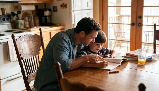 Father helping son with homework at sunlit kitchen table, family learning together