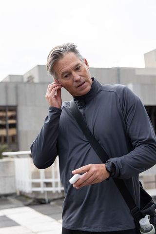 Mature man inserting wireless earbud on urban rooftop wearing smartwatch and crossbody bag