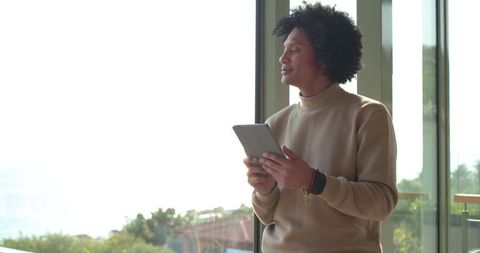 Contemplative African American man standing by window holding tablet, looking out