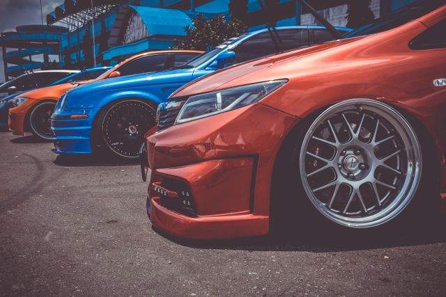 Colorful modified cars are lined up in a row, showcasing their custom wheels and body kits at an outdoor auto show. This can be used in promotional materials for automotive events, car enthusiast blogs, or advertisements for auto tuning and modification services.