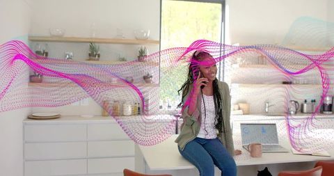 Smiling woman talking on phone at kitchen island, working on laptop with magenta data wave overlay