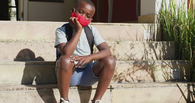Young boy sitting on outdoor steps wearing red face mask and backpack. Captures a thoughtful or waiting moment, perfect for themes like education, back to school, health precautions, and childhood experiences.