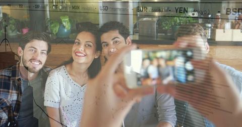 Smiling friends taking selfie at coffeehouse with currency ticker reflection and graphs