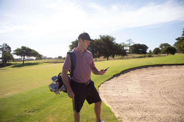 Caucasian male golfer standing near a sand trap on a sunny golf course, wearing a cap and golf clothes, carrying a golf bag and checking his smartphone. Ideal for use in advertisements for golf equipment, mobile apps, or promoting healthy outdoor lifestyles. Can also be used in articles about golfing, leisure activities, or technology in sports.