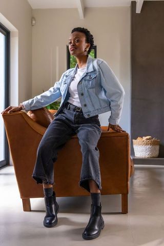 African american woman sitting on leather armchair wearing denim jacket and ankle boots, gazing