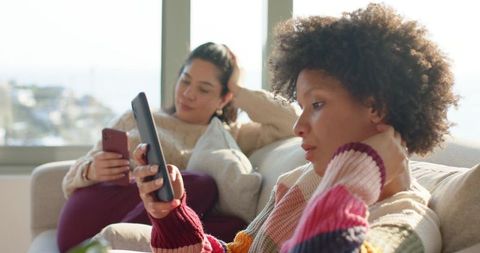 Two diverse female friends relaxing and scrolling smartphones on sofa wearing colorful knit sweaters