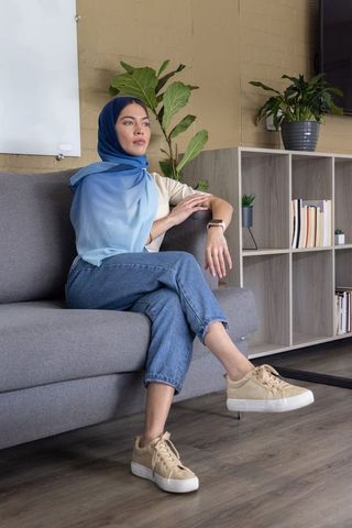Contemplative muslim woman sitting on gray sofa wearing blue ombre hijab and smartwatch