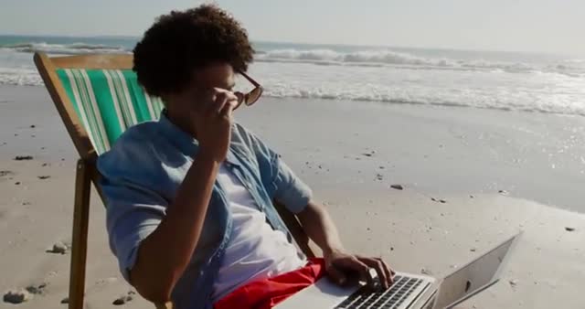 Young biracial man sits on a deckchair at the beach, using a laptop while enjoying sunny weather. Ideal for themes on remote work, digital nomad lifestyle, leisure, technology, and summer vacations.