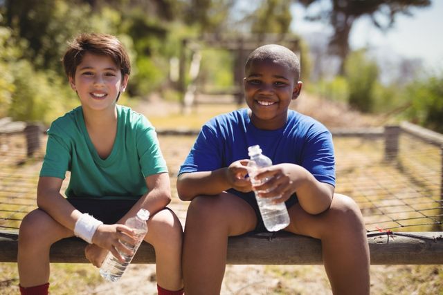 Two boys are sitting and smiling while holding water bottles after completing an obstacle course workout. This image can be used for promoting children's fitness programs, summer camps, teamwork activities, and healthy lifestyle campaigns.
