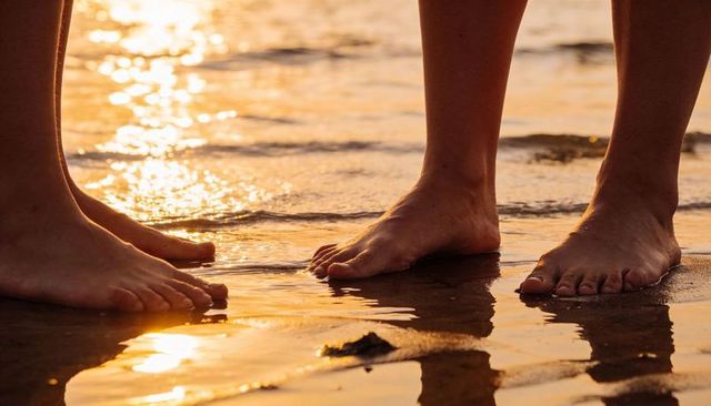 Barefoot couple standing and wading on sunlit wet sand at sunset shoreline golden glow