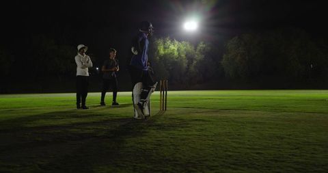 Night cricket batter standing at crease under stadium floodlight wearing helmet and pads