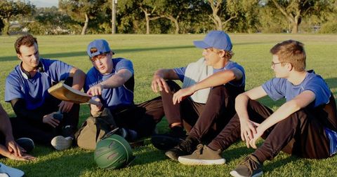 Young diverse cricket teammates resting on grass after practice passing bat and laughing