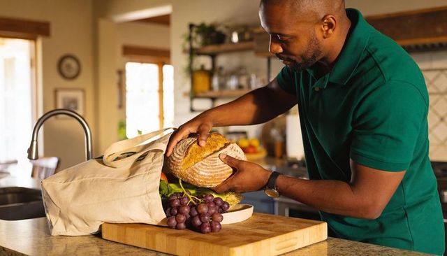 Man unpacking fresh groceries: placing artisan loaf on cutting board in sunlit kitchen