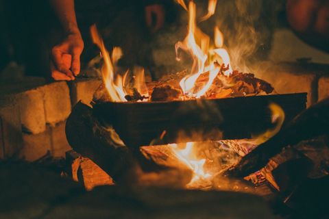 Campfire flames crackling over burning logs, hand tending ember for cozy night gathering