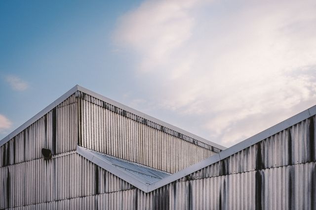 Industrial building showcasing corrugated metal roof against blue sky with clouds. Ideal for illustrating themes related to industrial environments, modern architecture, construction materials, and urban landscapes. Suitable for use in brochures, websites, and educational materials focused on architecture and construction.