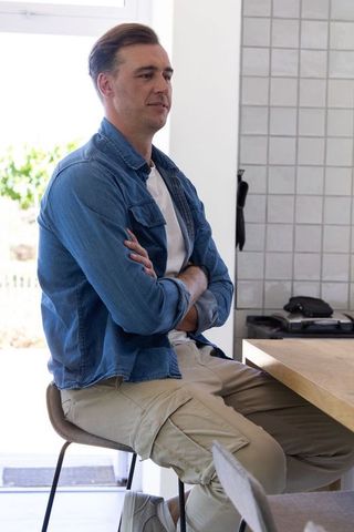 Middle-aged man sitting on stool in sunlit kitchen wearing denim shirt and cargo pants