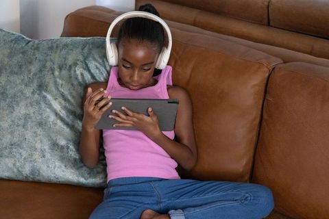 African american girl using tablet on brown couch wearing headphones and concentrating