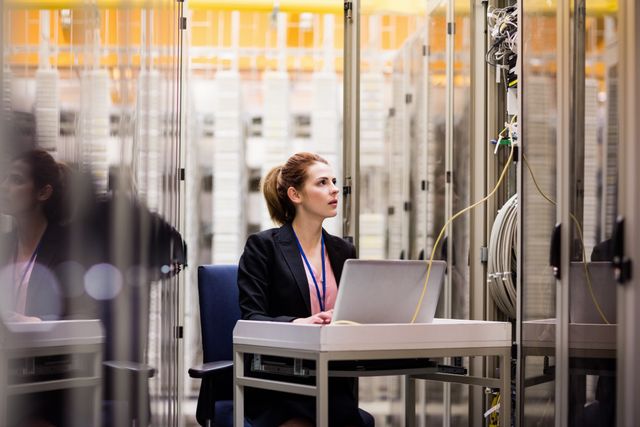Female technician working on a laptop while analyzing server connections in a data center. Ideal for use in articles or advertisements related to technology, IT services, cybersecurity, and network management. Can also be used for illustrating modern business environments and professional IT roles.