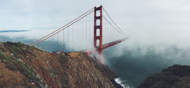 Photograph of the Golden Gate Bridge partially shrouded in fog over the San Francisco Bay. Red skyscrapers extending from scenic cliffs into the water create a mystical, atmospheric backdrop. Ideal for capturing the essence of San Francisco, usable in tourism materials, travel blogs, scenic calendars, and cultural presentations.