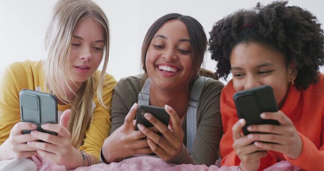 Three teenage girls laying on bed, spending time together, using smartphones. They are smiling, indicating a joyful and engaging moment. Suitable for use in topics like technology, social media influence, friendship, and teenage lifestyle. Ideal for illustrating youthful connections and digital communication.