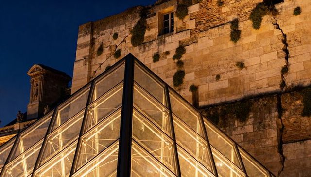 Glowing glass-and-steel pyramid illuminating historic stone wall courtyard at night