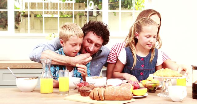 Family having breakfast together in a bright kitchen. Parents enjoying time with their children. Fresh fruit, juice, and bread on table. Ideal for advertisements, family lifestyle blogs, or articles on healthy living.