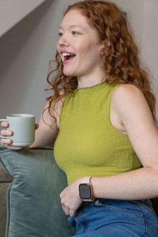 Laughing redhead woman holding mug on sofa wearing lime textured top and smartwatch