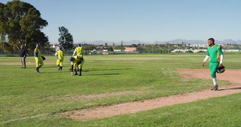 Baseball player walking base path holding helmet with teammates warming up on sunny field